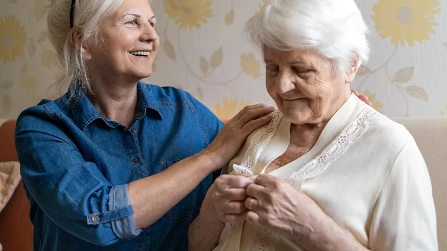 Eine Frau hilft einer Seniorin in einem Wohnzimmer beim Ankleiden.