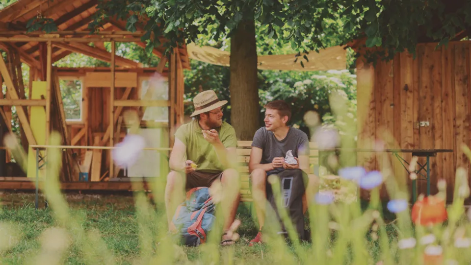 Zwei junge Männer sitzen auf einer Bank in einem Garten und essen Brote.
