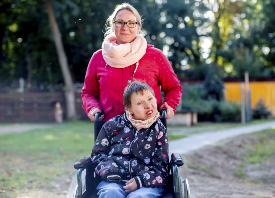 Eine Frau schiebt eine junge Frau mit Behinderung, die im Rollstuhl sitzt.