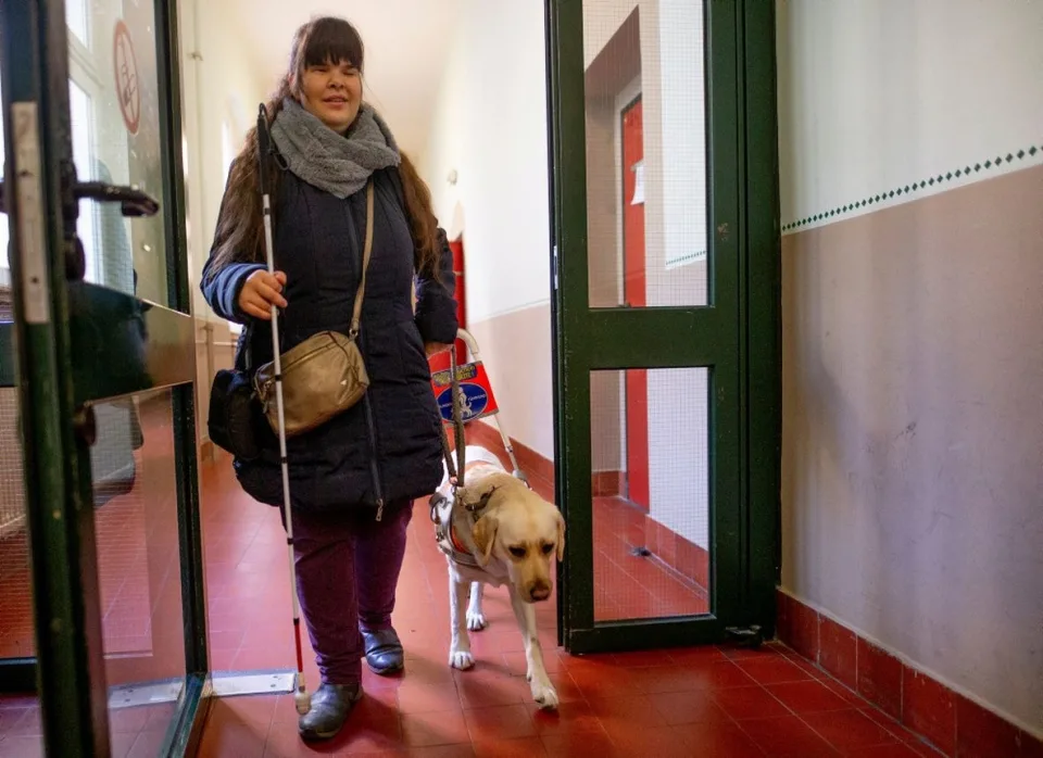 Eine blinde Frau geht mit einem Assistenzhund durch eine Glastür in einem öffentlichen Gebäude.