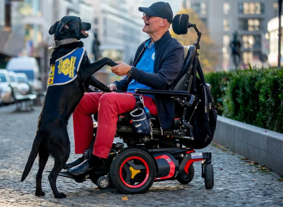 Ein Mann im Rollstuhl hält einen Assistenzhund an den Vorderbeinen fest. Mann und Hund sind auf einer Augenhöhe und gucken sich an.