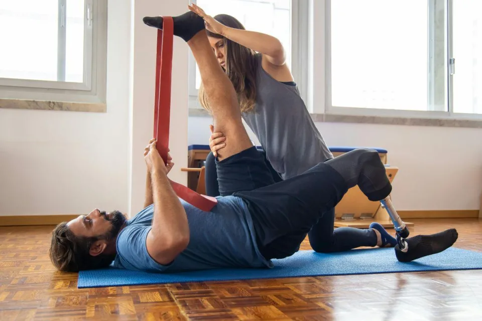 Ein Mann mit einer Beinprothese liegt auf einer Gymnasikmatte und macht eine Bewegungsübung. Eine Therapeutin unterstützt ihn dabei.