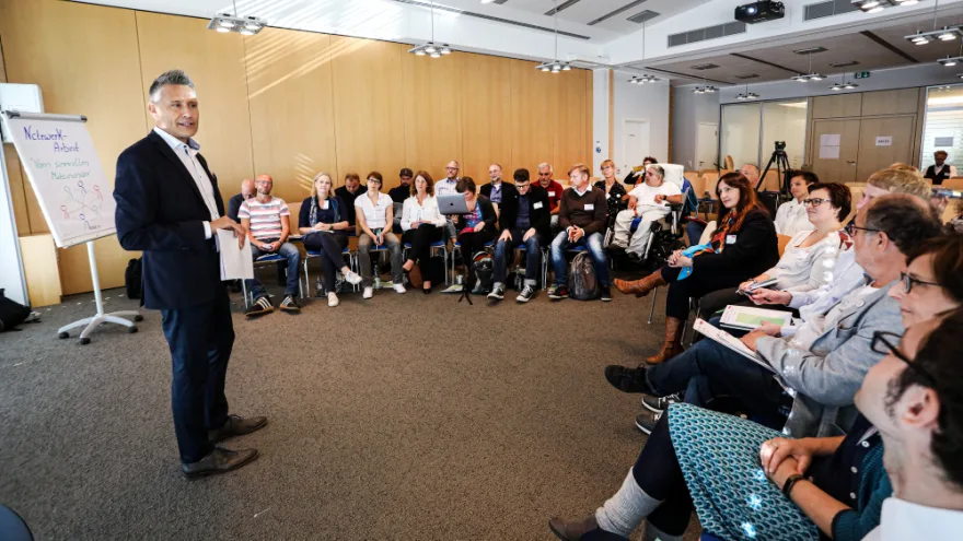 Ein Workshop zum Thema Netzwerk-Arbeit. Eine Gruppe von Menschen sitzt in einem Raum. Vor ihr steht ein Mann und erklärt etwas.